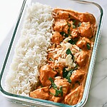 Creamy coconut chicken served with white rice in a glass container topped with toasted coconut and green herbs