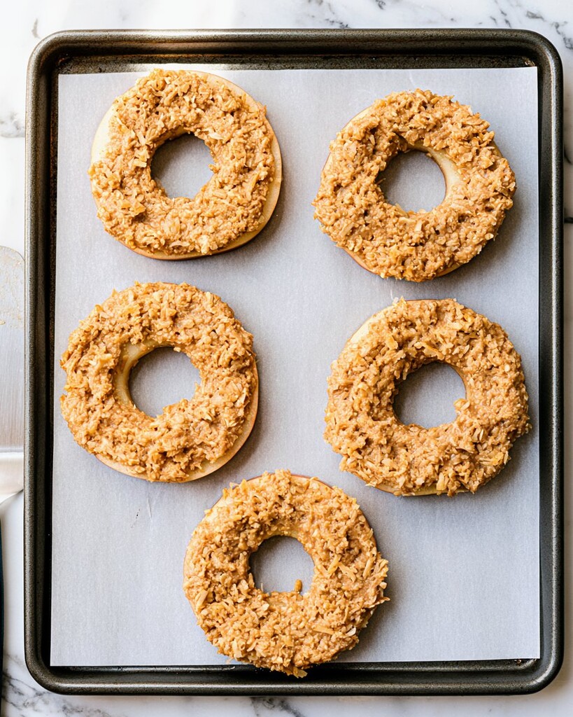 Thick coconut mixture spread evenly over apple rings on a parchment lined baking sheet