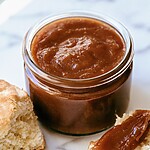 Dark brown vanilla bean apple butter in a glass jar beside smeared biscuits