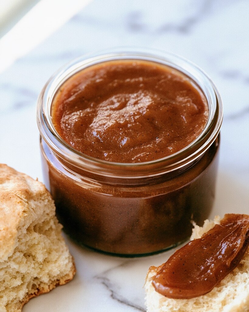 Dark brown vanilla bean apple butter in a glass jar beside smeared biscuits
