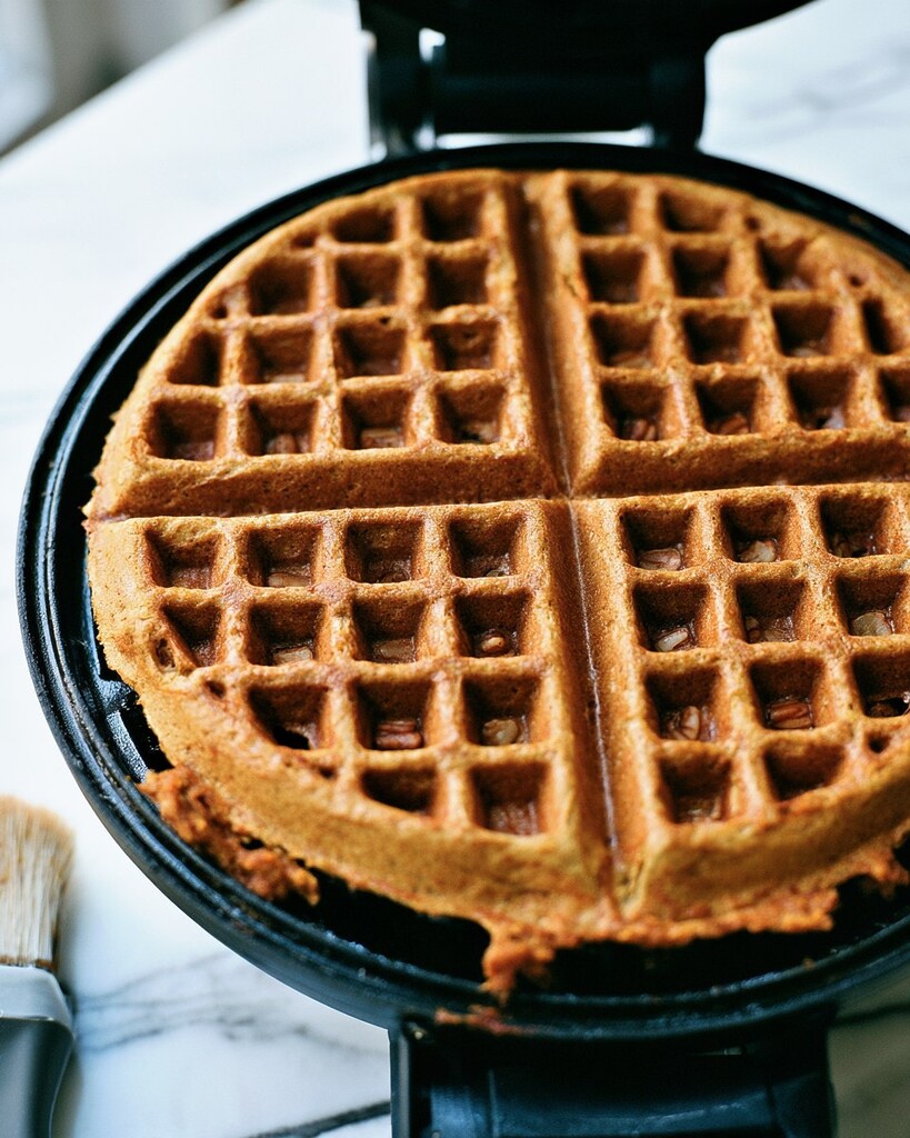 Pumpkin oat waffle cooking inside a waffle maker with toasted pecans on the golden surface