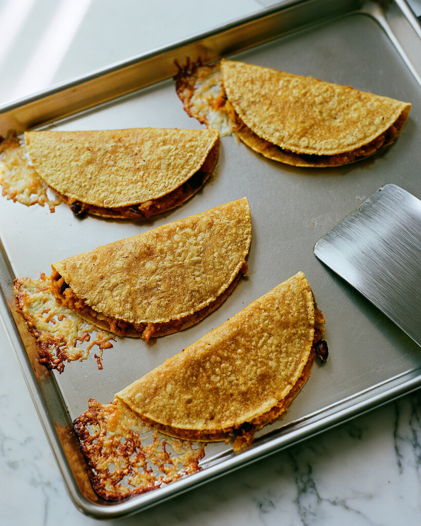 Folded pumpkin and black bean tacos baking on a metal sheet pan with melted cheese bubbling out the edges