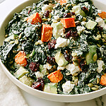 Brightly lit bowl of sweet potato and kale salad topped with diced avocado, goat cheese, dried cherries, and pepitas