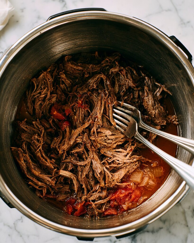 Two forks shredding salsa-braised beef directly in the dark juices inside a pressure cooker