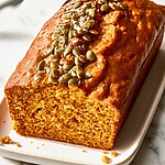A whole pumpkin bread with maple-candied pepitas showing a golden crust and orange crumb on a serving plate