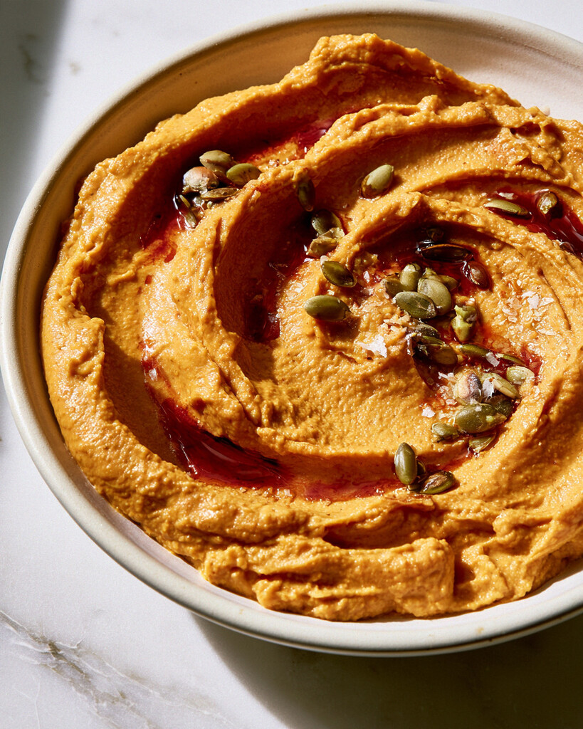Spreading orange pumpkin hummus into a shallow ceramic bowl with swirling adobo and pumpkin seeds