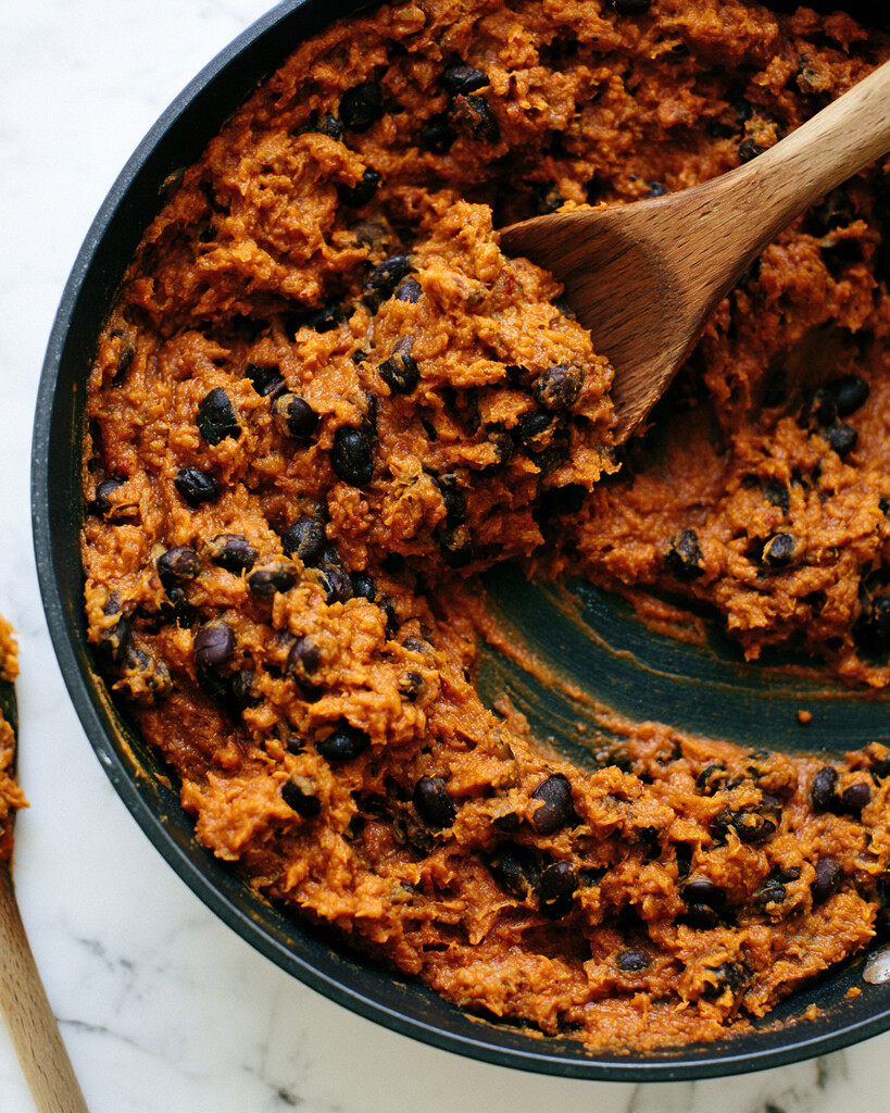 Thick spiced pumpkin and black bean taco filling cooking in a black skillet with a wooden spoon
