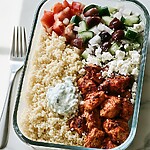 Glass container holding a serving of spiced chicken, couscous, chopped vegetable salad, and a dollop of tzatziki