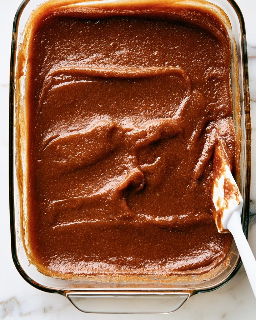 Thick, dark brown vanilla bean apple butter in a glass baking dish with a silicone spoon resting inside