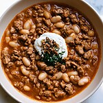 Smoky Chipotle Pumpkin Turkey Chili (Ready in 45 Minutes) Overhead view of thick orange turkey chili garnished with pepitas, crema, and fresh herbs in a white bowl