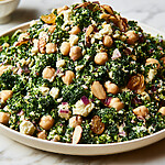 A dense chopped broccoli and chickpea salad with feta, almonds, and golden raisins in a light serving bowl.