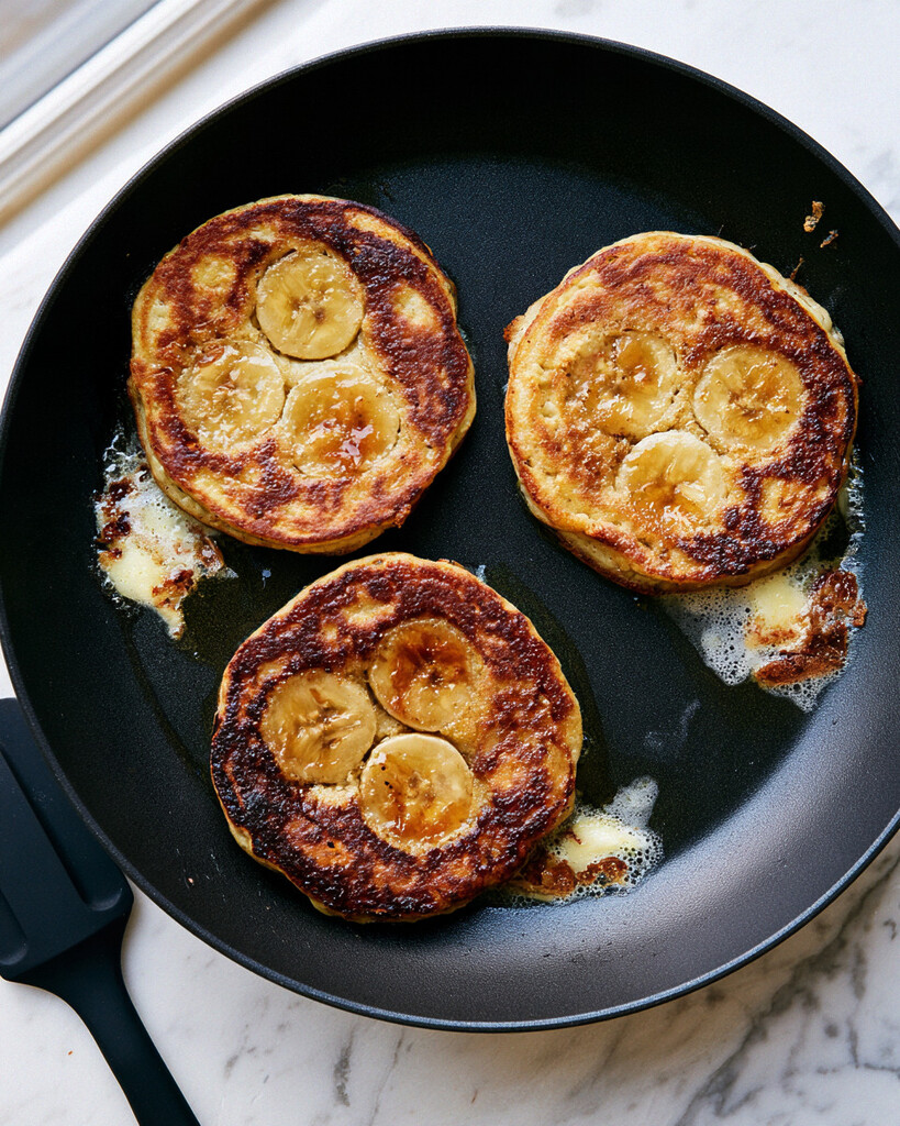Pancake batter cooking in a nonstick skillet with sliced bananas pressed into the tops