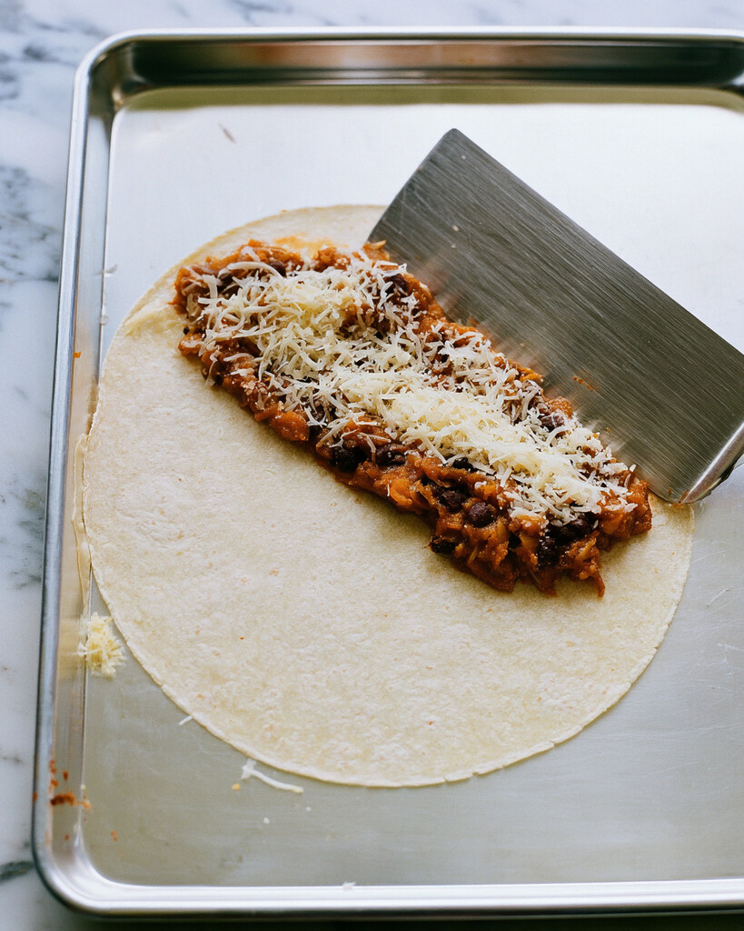 Spiced pumpkin and black bean filling and grated cheese portioned onto an oiled corn tortilla half on a bare baking sheet