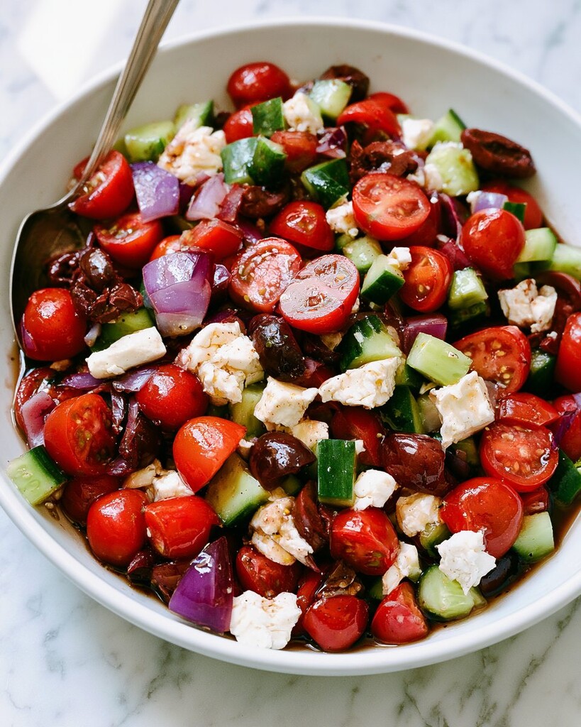 White bowl filled with chopped cherry tomatoes, diced cucumber, red onions, olives, and crumbled feta cheese