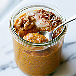 Small glass jar filled with thick orange pumpkin chia pudding topped with white cream, pecans, and a spoon resting inside