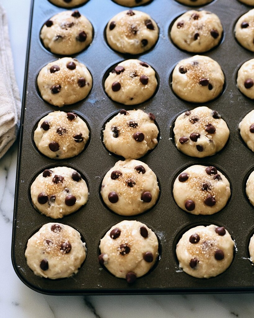 Mini muffin pan filled with thick batter and generously topped with chocolate chips and raw sugar