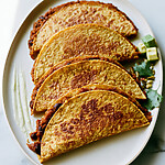 Crispy baked pumpkin tacos arranged on an oval plate with lime wedges, diced avocado, and fresh cilantro