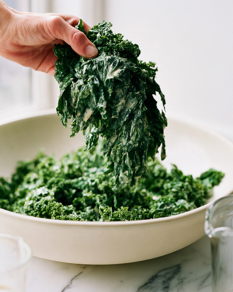 Two hands massaging creamy tahini dressing into dark green kale leaves in a large bowl