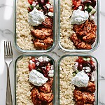Greek Chicken Couscous Meal Prep Bowls That Won't Get Soggy Four glass meal prep containers lined up, each filled with fluffy couscous, spiced roasted chicken, chopped horiatiki salad, and thick tzatziki