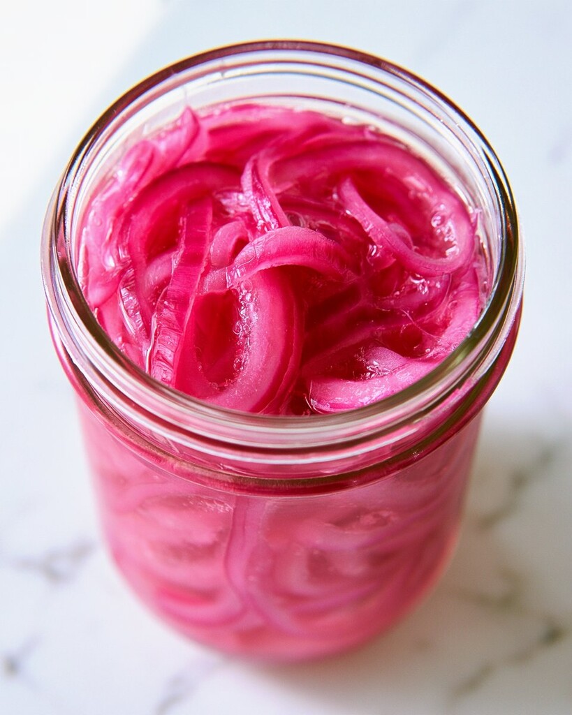Sliced red onions submerged in a bright pink pickling liquid in a glass jar