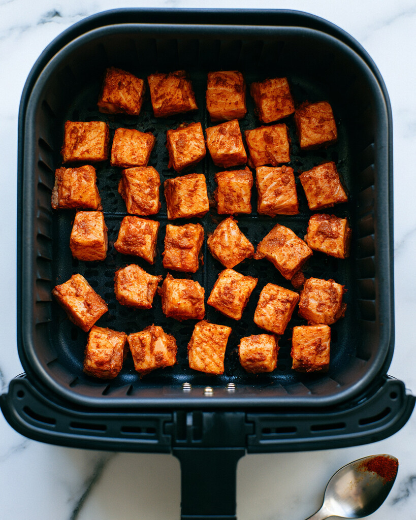 Seasoned salmon cubes arranged in a single layer inside an air fryer basket with space between pieces