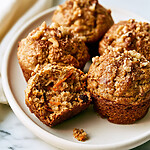 Golden baked carrot banana muffins on a plate with one broken open to show the moist carrot-flecked crumb