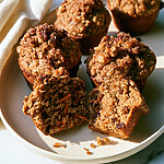 Golden carrot banana muffins on a white plate with a walnut-sugar crust and two halves showing carrots and raisins