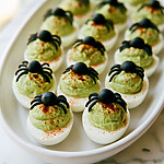 Sliced black olive halves arranged as spider bodies and legs over green deviled egg filling sprinkled with red paprika