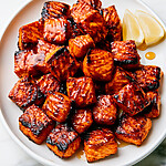 Crisp blackened salmon bites with a glossy hot honey glaze stacked on a round white plate beside fresh lemon wedges